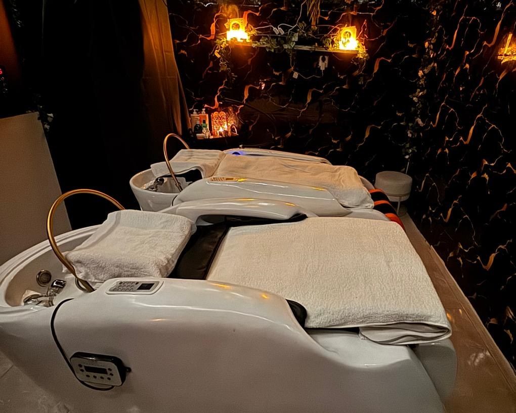 De head spa-kamer van Sadaf Beauty Salon: twee witte head spa-bedden, kaarslicht en groen aan een donkere wand